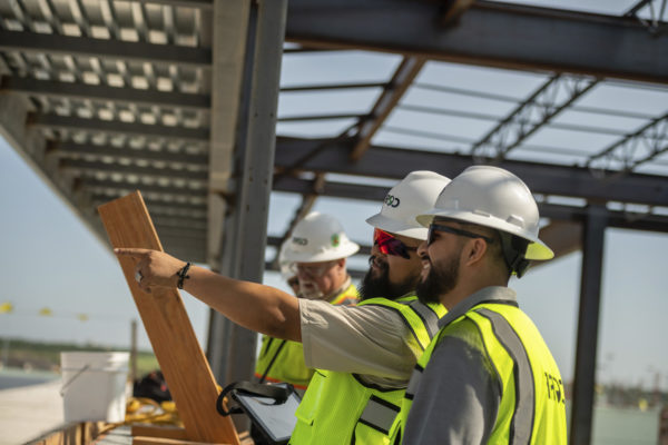 Three men in white hardhats and safety vests in discussion on a construction site.