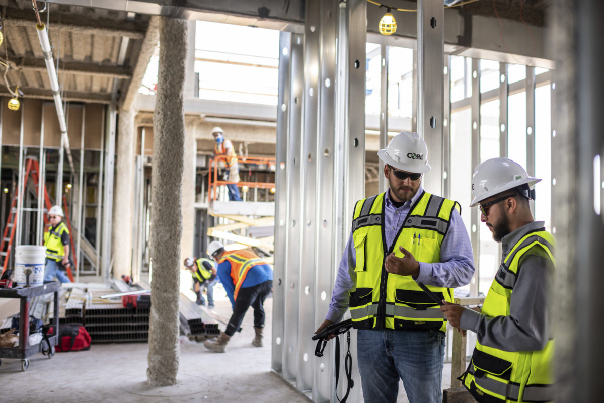 Two construction workers with hard hats and safety vests looking at a tablet and conversing.